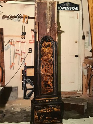 Antique wooden grandfather clock with intricate Asian artwork in a workshop.