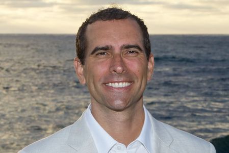 Smiling man in a light suit with ocean background at sunset.