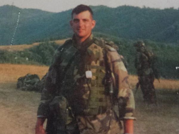Soldier in camouflage gear standing in a field with another soldier in the background.