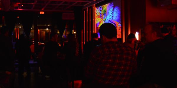 Patrons watch comedy in a neon-red lit room. A video of twisting colors plays on a projector