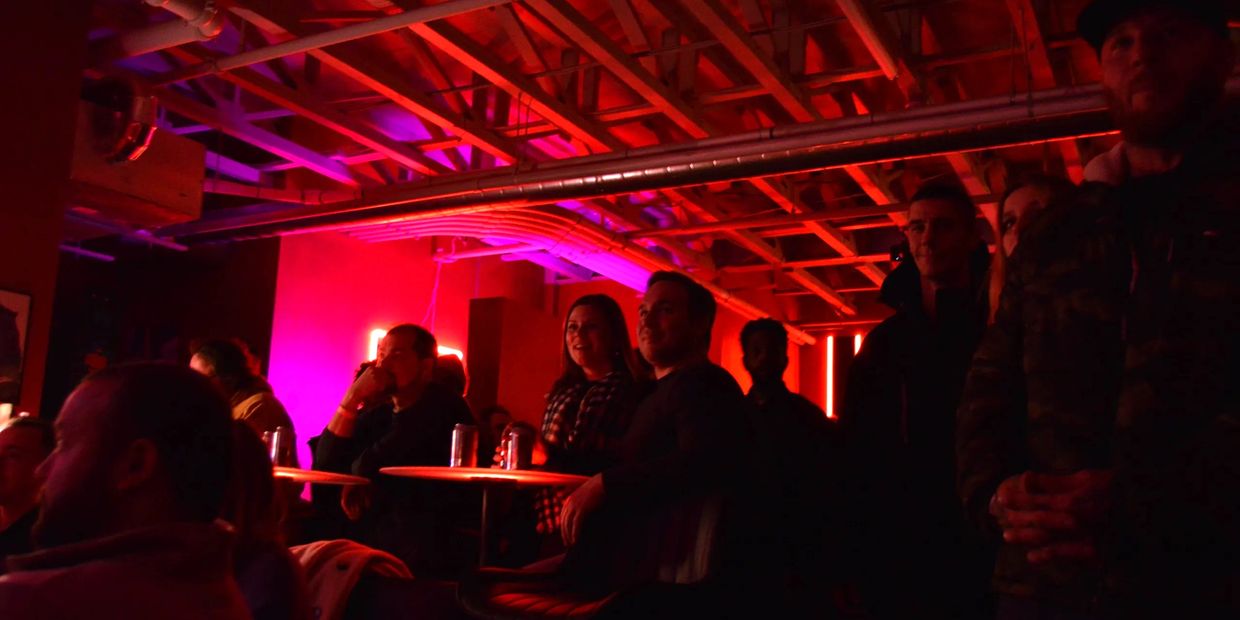Patrons laugh at jokes told by a comedian under red lights while sipping canned beverages