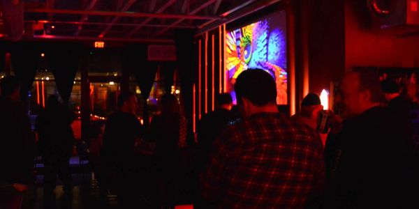 Patrons watch comedy in a neon-red lit room. A video of twisting colors plays on a projector