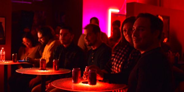 A group of people sit at neon red hightop tables drinking from cans and smiling