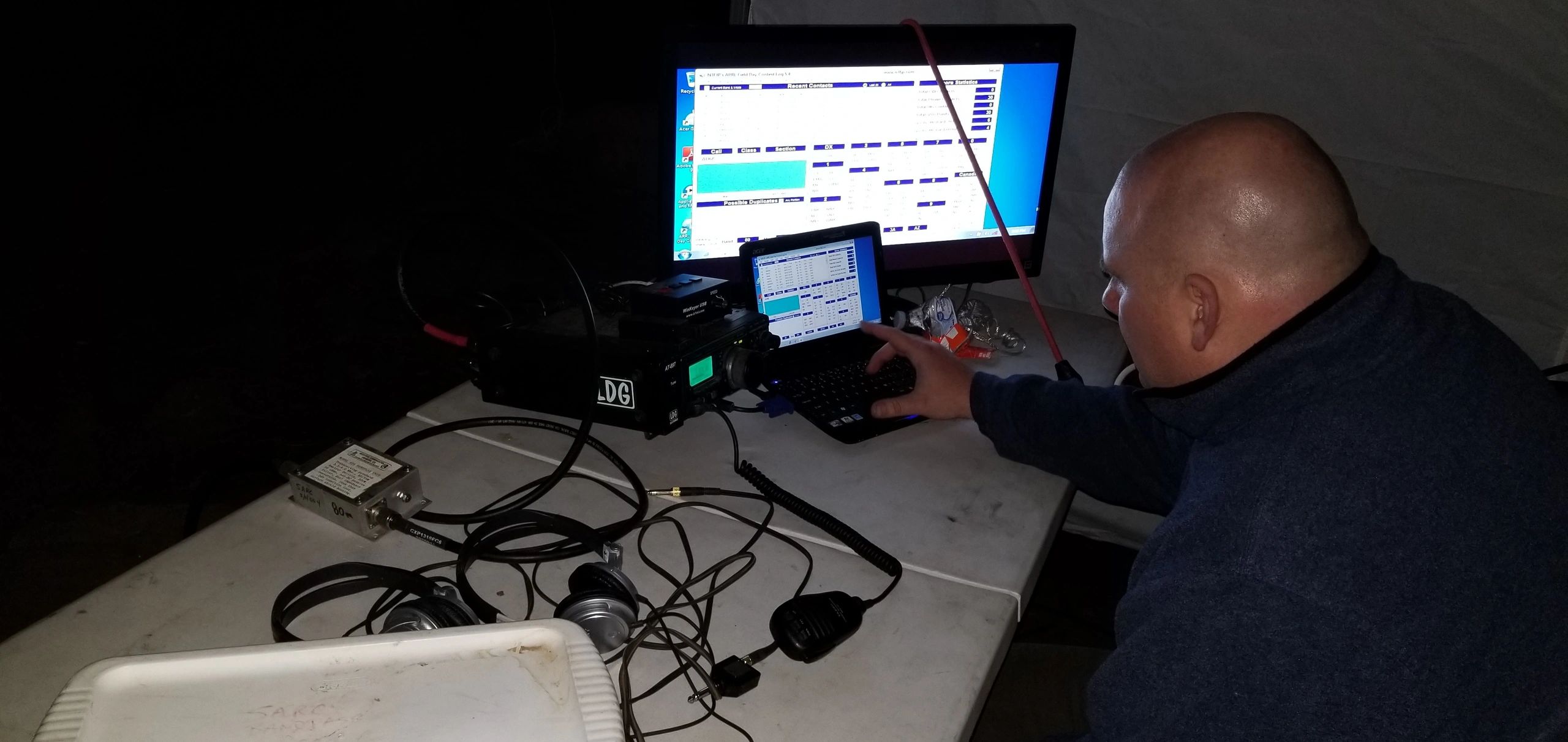 Man operating radio communication equipment at night on a cluttered table.