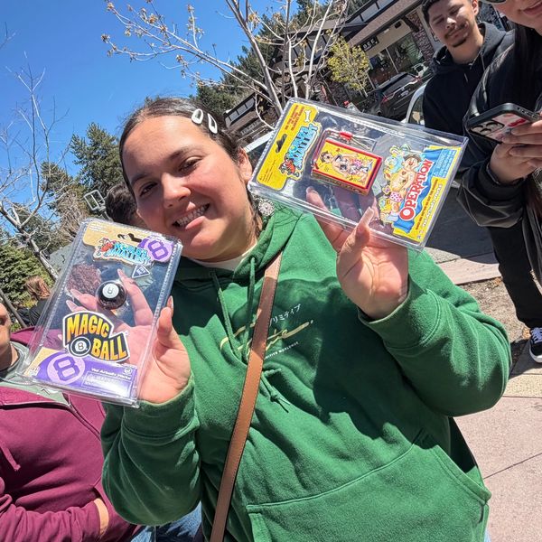 A smiling woman in a green hoodie holds up two retro handheld games outdoors on a sunny day.