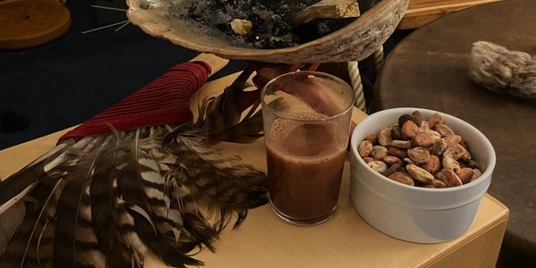 A ceremonial setup with feathers, a shell bowl with ashes, cacao beans, and a glass of cacao drink.