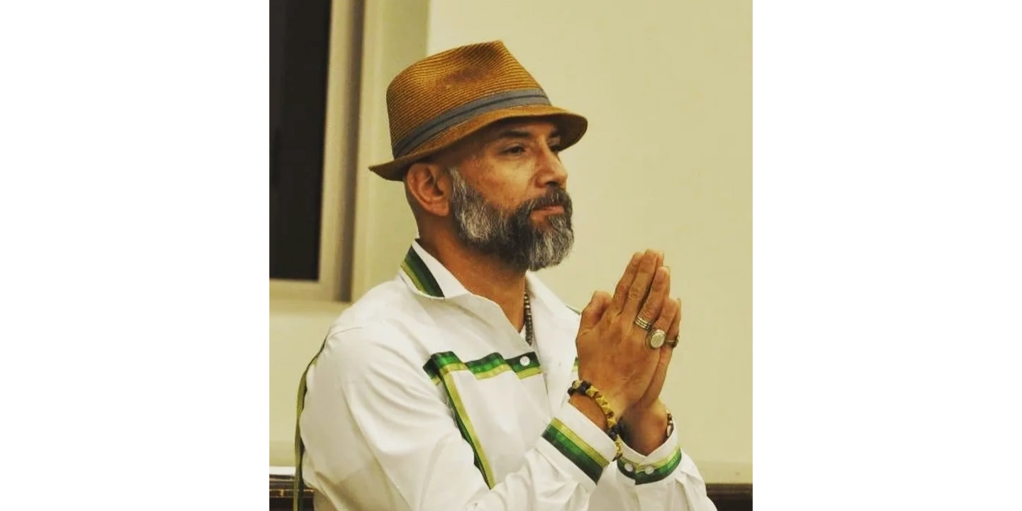 Man with a beard and hat sitting thoughtfully with hands clasped.