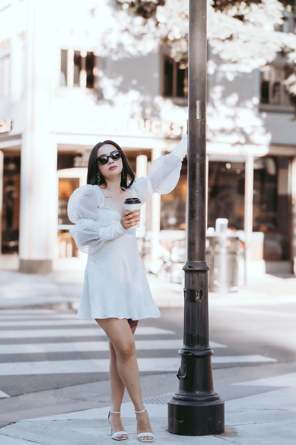 Woman in a white dress holding coffee and leaning on a street lamp.