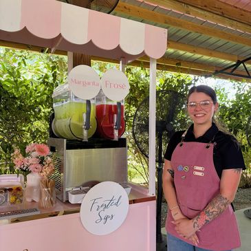 Woman in pink apron stands by a pastel pink drink station with margarita and frosé slushies.