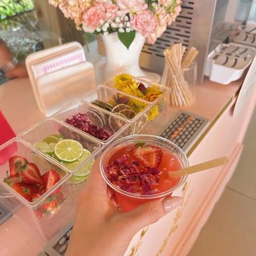 Hand holding a floral-infused drink with fresh fruit and flower garnishes in the background.