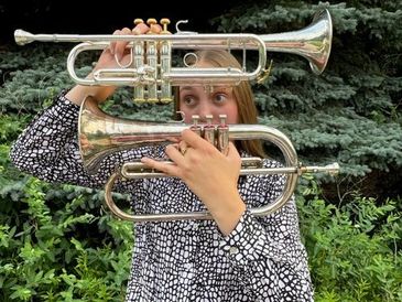 Person holding two brass instruments in front of their face outdoors.