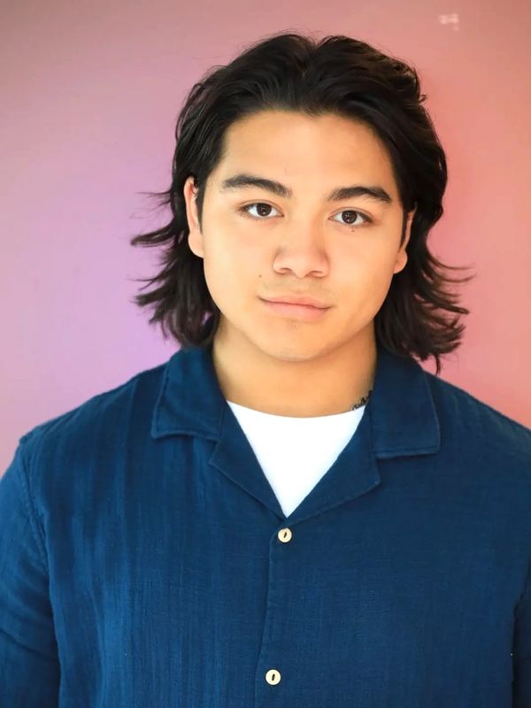 Young man with medium-length hair wearing a navy blue shirt against a pink backdrop.