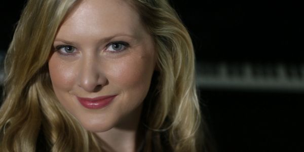 Close-up of a smiling blonde woman with a blurred piano in the background.