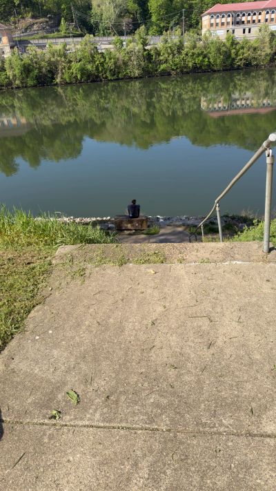 A person sitting by a calm river surrounded by greenery.