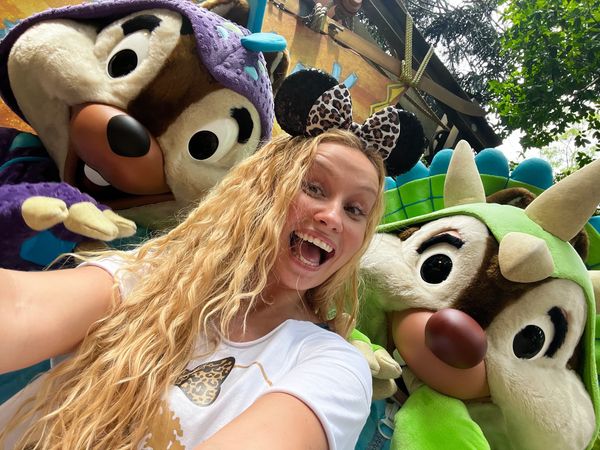 A woman happily poses with two Disney characters in colorful costumes.