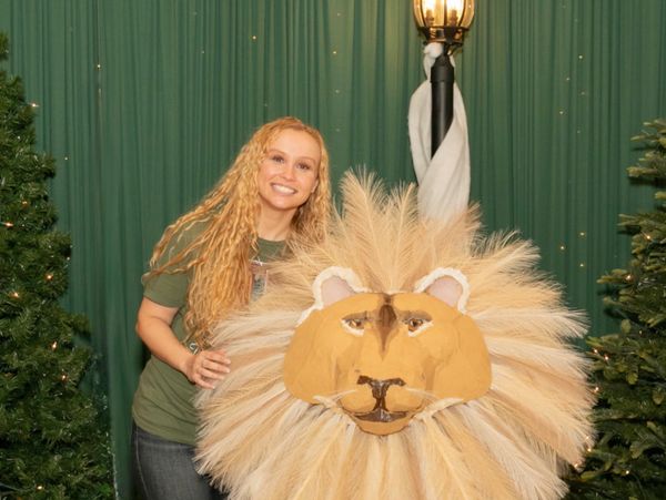A smiling woman poses next to a large lion head prop with a fluffy mane.