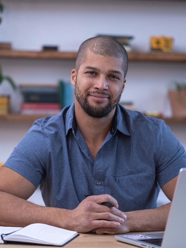 Man in blue shirt sitting at desk with notebook and laptop.
