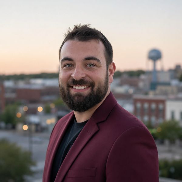 Smiling man with beard wearing a maroon blazer outdoors.