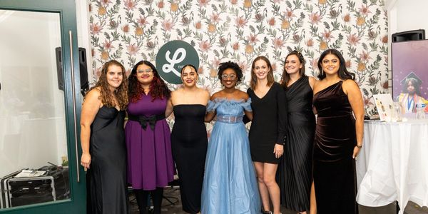 Seven women dressed elegantly posing together at an indoor event.
