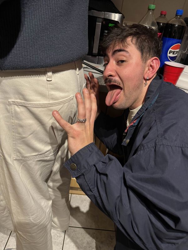 Man playfully sticks out his tongue near another person's back pocket indoors.