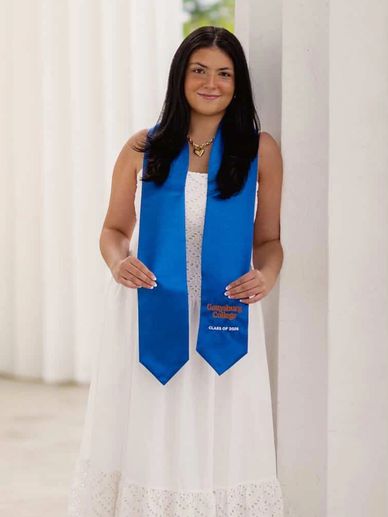Young woman in white dress with blue graduation stole posing indoors.