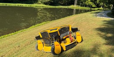 A yellow robotic lawn mower on a grassy slope near a pond and trees.