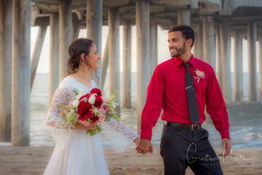 Couple holding hands on beach under pier, dressed formally.