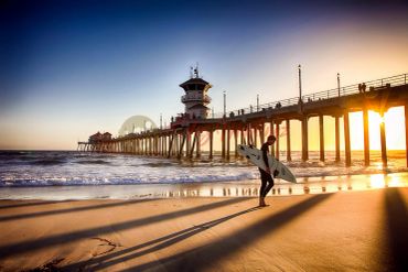 Surfer walking on the beach near a pier during sunset.