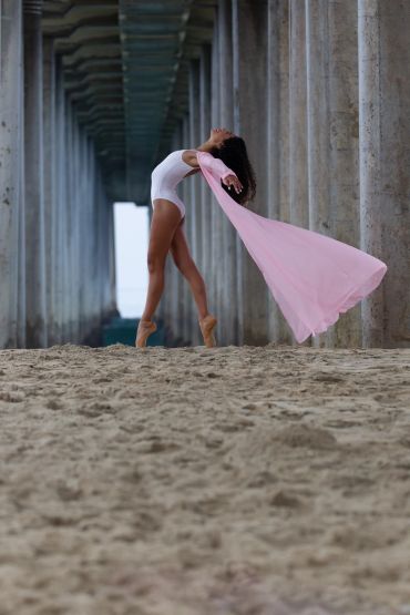 Ballet dancer in white bodysuit and pink fabric poses gracefully on sand.