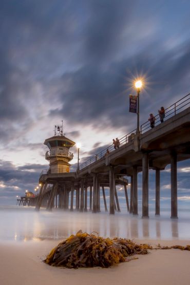 Pier at sunset with cloudy sky and seaweed on beach.