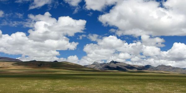 Vast grassy plain under a bright blue sky with fluffy clouds and distant mountains.