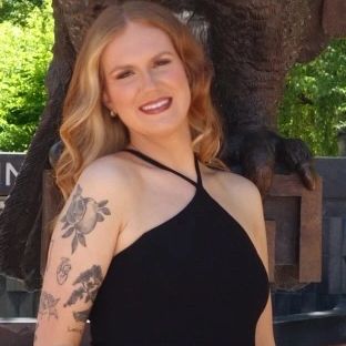 Smiling woman with blonde hair and floral tattoos, wearing a black halter dress.