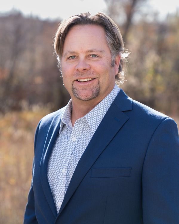 Smiling man in a blue blazer outdoors with a patterned shirt.