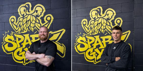 Two men posing in front of a Spartan gym logo on a black wall.