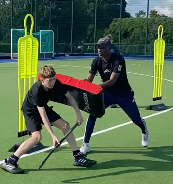A coach trains a young hockey player using a padded shield on the field.