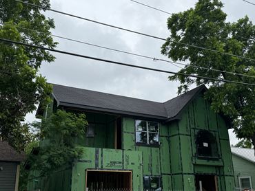 A partially constructed two-story house with green sheathing and trees around.