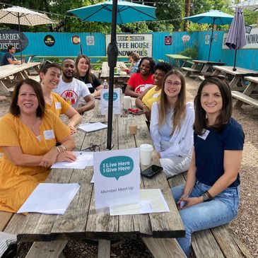 A group of diverse people smiling at a community meet-up outdoors at a picnic table.