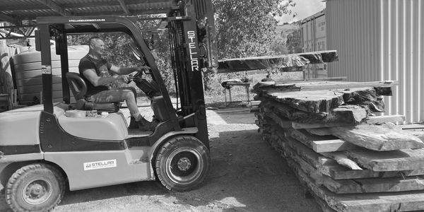 Man operating a forklift to move large wooden slabs outdoors.