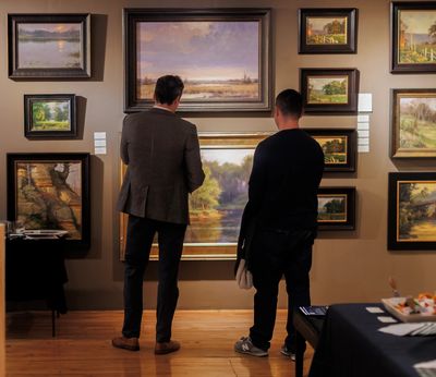 Two people stand side by side viewing a framed landscape painting on a wall of framed artwork at Gal