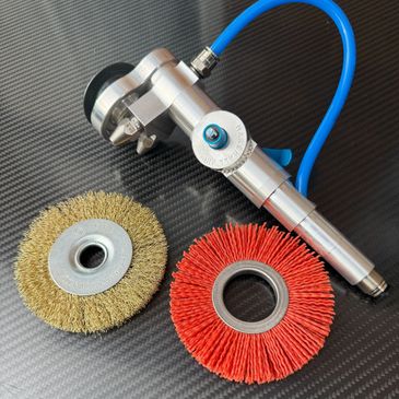Air-powered rotary tool with wire and nylon brush attachments on carbon fiber surface.