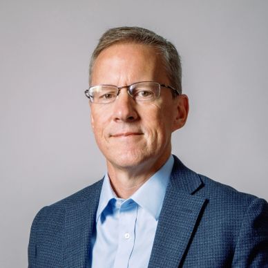 Man wearing glasses and a blue blazer against a gray background.