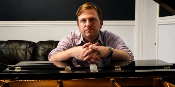 Man leaning on a grand piano in a cozy room.
