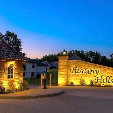 Illuminated Tuscany Hills entrance at dusk with stone walls and a guardhouse.