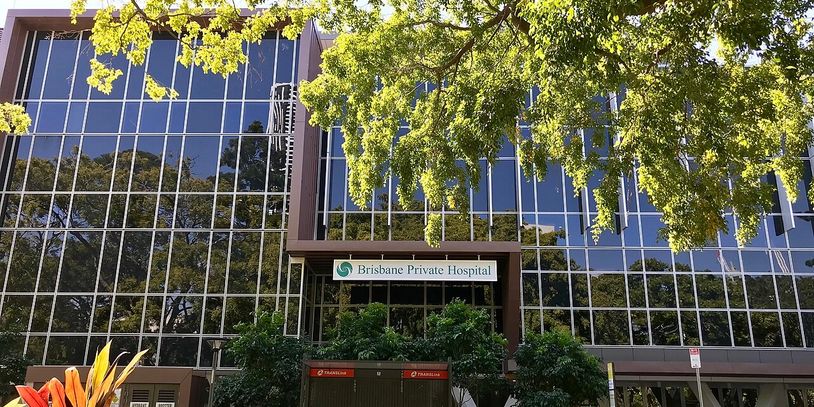 Modern glass building of Brisbane Private Hospital surrounded by greenery.