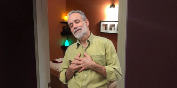 A man in a green shirt stands with hands on chest, eyes closed, smiling peacefully.
