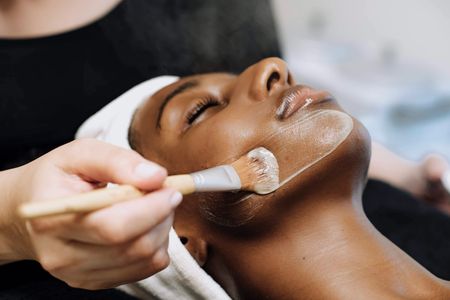 A woman receives a facial treatment with a brush applying a cream mask.