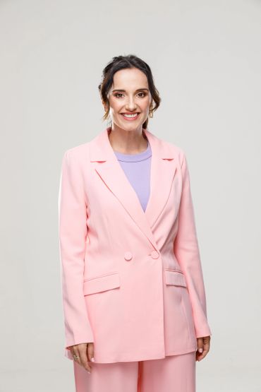 Smiling woman in a pastel pink suit and lavender shirt against a plain backdrop.