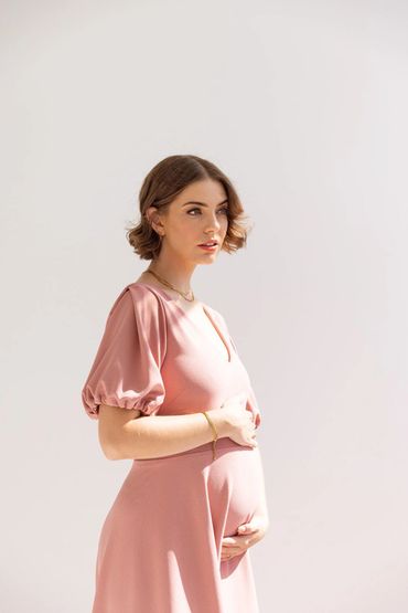 Pregnant woman in a pink dress gently cradles her belly, looking thoughtful.