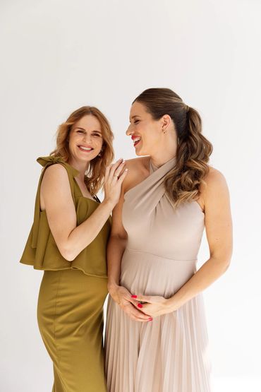 Two women in elegant dresses share a joyful moment against a plain background.