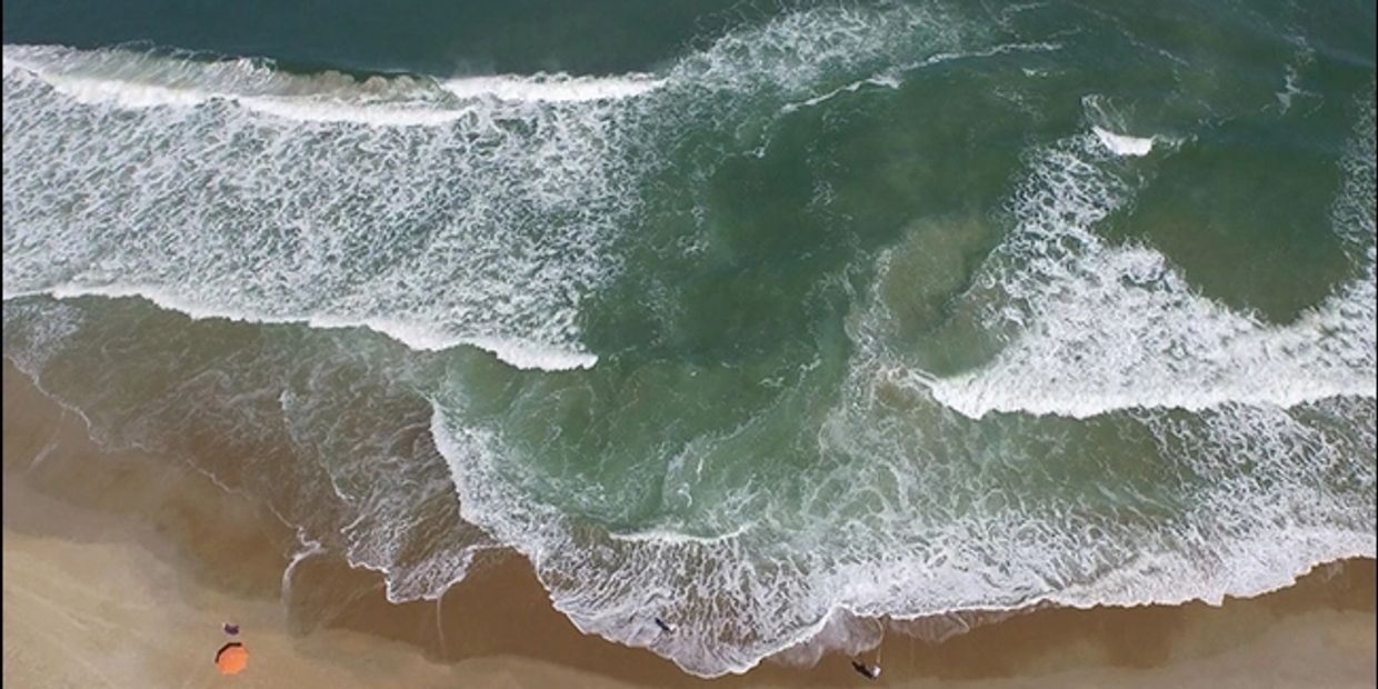 Aerial view of waves crashing on a sandy beach with a few people and an orange umbrella.